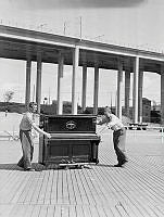 Skanstull. Förberedelser för kommunal parkdans i Eriksdalslunden. Två män rullar ett piano, Skanstullsbron i fonden.