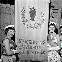 Hantverkargatan 1. Stockholms högskola blev Stockholms universitet. Invigning av det nya universitetet firas i Stadshuset. Två flickor i studentmössa och långklänningar står vid studentkårens fana.