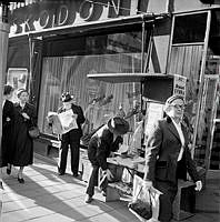 Tidningförsäljning på gatan. Drottninggatan 65. Tidningsförsäljning utanför Kembels skor. En tidningsförsäljare har slagit upp ett bärbart tidningsstånd.