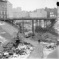 Vy norrut från Klarabergsgatan över rivningar och tunnelbanebygge kring platsen för blivande Sergels Torg. Mäster Samuelsgatan går via tillfällig bro.