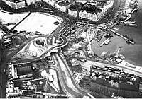 Flygbild över Slussen. Västra delen av Slussenkarusellen under byggnad. Den östra delen ännu ej påbörjad. Till vänster syns Stadsmuseet. till höger Stadsgården och kvarteret Tranbodarne.
